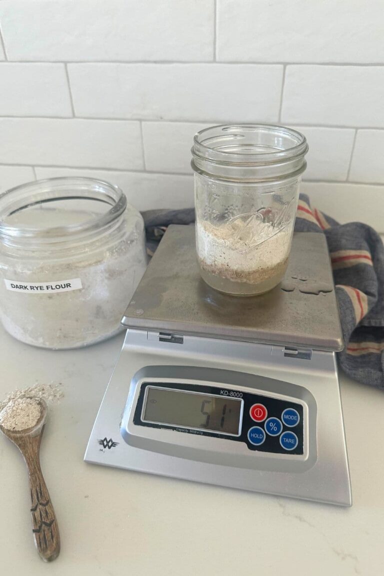 A photo of a 16oz mason jar sitting on a scale. The jar is filled with water and dark rye flour to create a rye flour sourdough starter. You can also see a large jar of rye flour in the background and a wooden spoon with rye flour on it next to the scale.