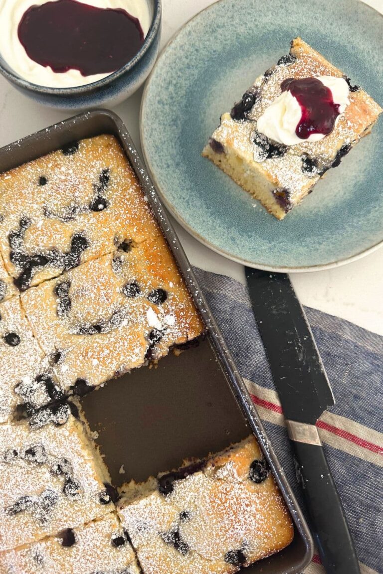 A photo of a tray of sourdough sheet pan pancakes with a piece missing from the tray. The piece of pancake is sitting on a blue plate to the right of the tray, garnished with vanilla yogurt and berry coulis.