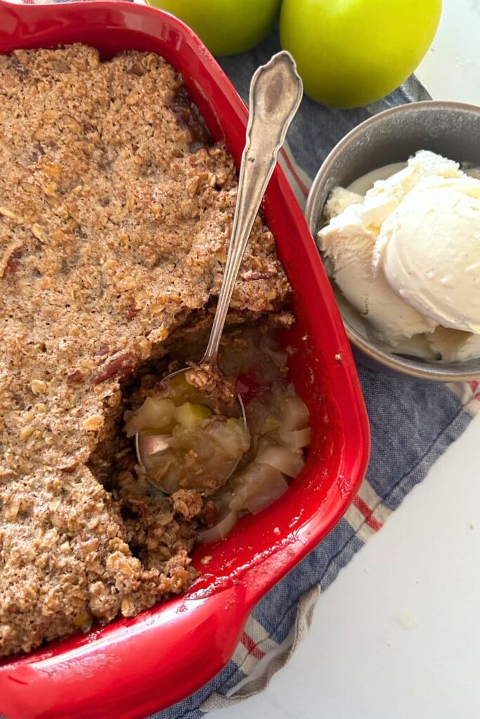 Sourdough apple crisp baked in a red baking dish. There are green apples and a bowl of vanilla icecream sitting to the right of the red baking dish. Some of the sourdough apple crisp has been scooped out using a vintage silver spoon.