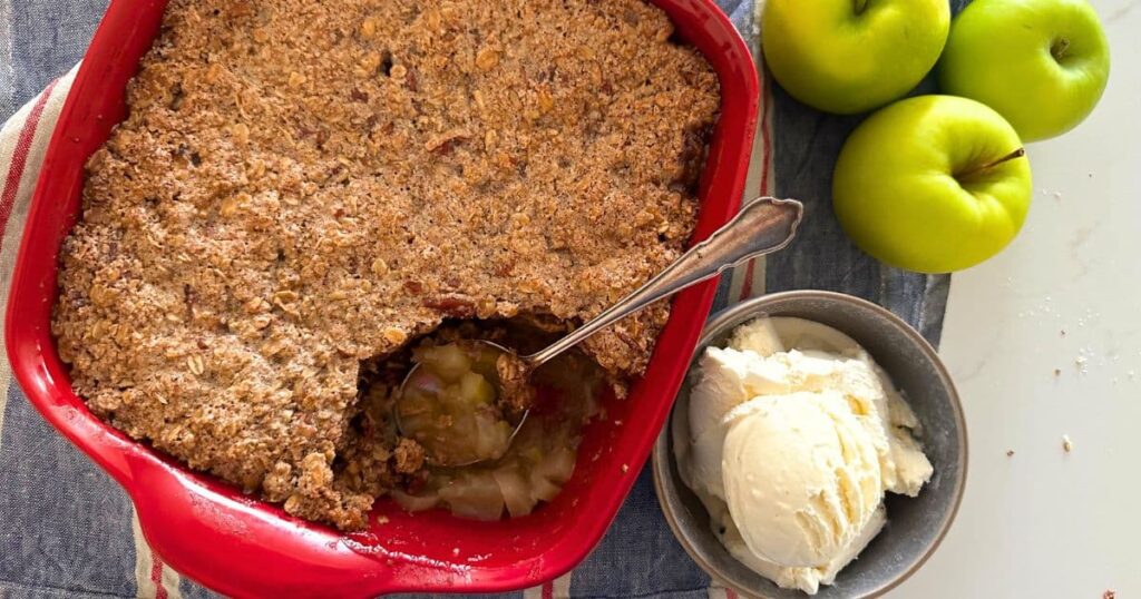 Sourdough apple crisp baked in square red baking dish. A piece of the apple crisp has been removed and placed into a bowl with vanilla ice cream and there are 3 green apples to the right of the baking dish.