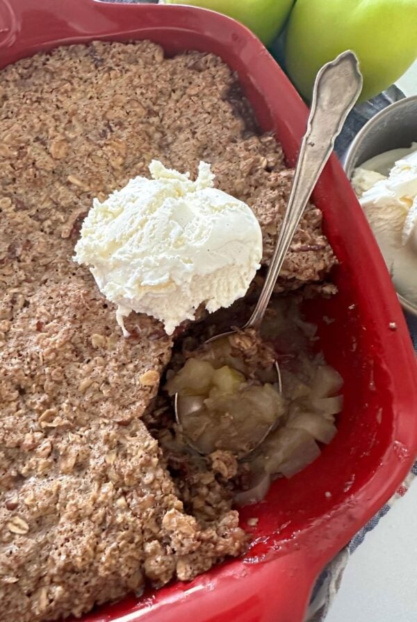 Sourdough apple crisp baked in a red baking dish. There are green apples and a bowl of vanilla icecream sitting to the right of the red baking dish. Some of the sourdough apple crisp has been scooped out using a vintage silver spoon.