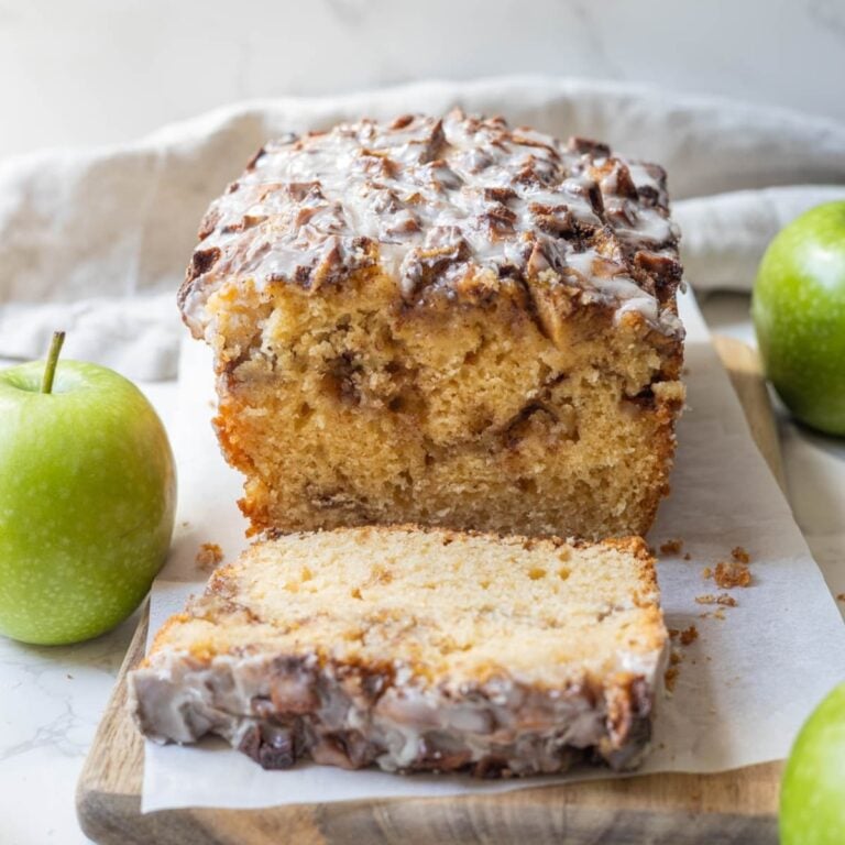 A loaf of sourdough discard apple fritter bread sliced open so you can see the cinnamony apple ribbon through the centre. There are some green apples displayed in the photo also.