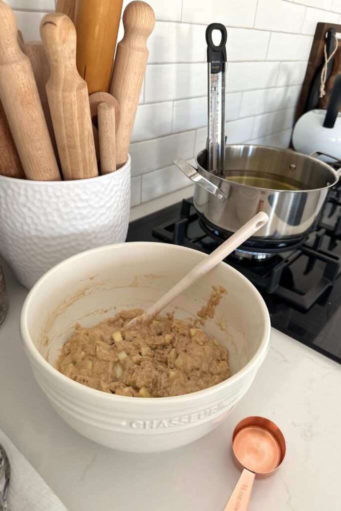 A cream colored mixing bowl containing sourdough apple fritter batter sitting on a white stone benchtop in a kitchen. You can see a stainless steel pot of oil sitting on the range ready to fry the fritters.