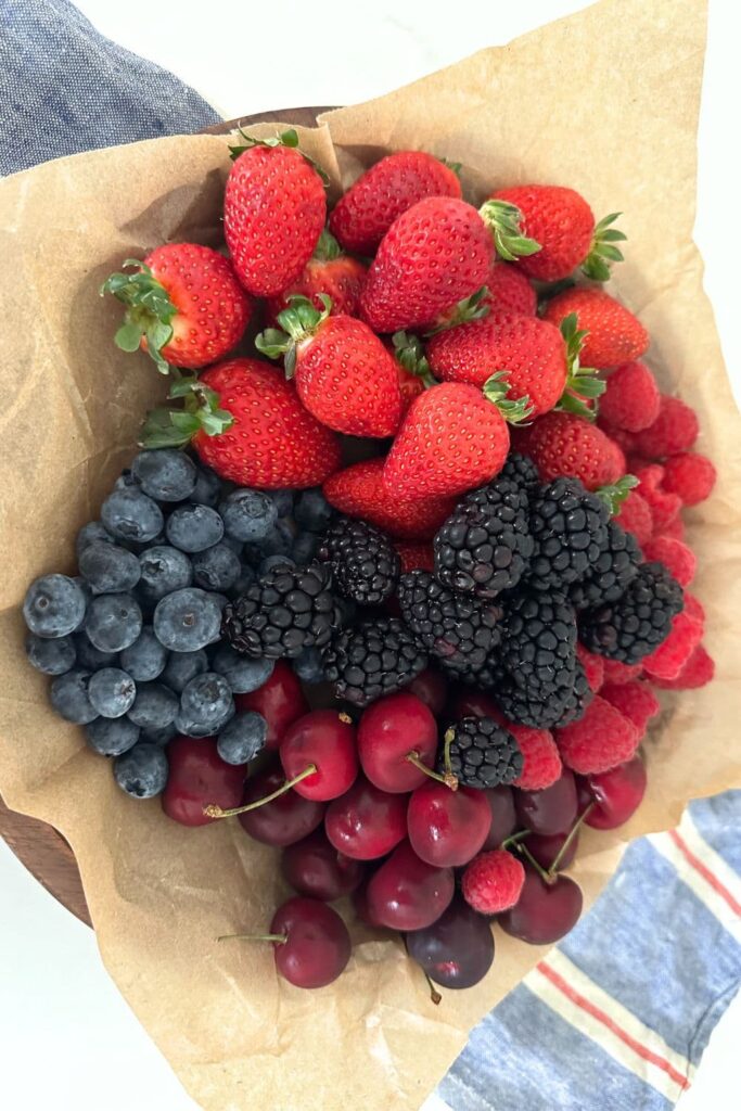 A selection of summer berries arranged on a sheet of parchment paper. There are strawberries, blueberries, cherries, raspberries and blackberries.