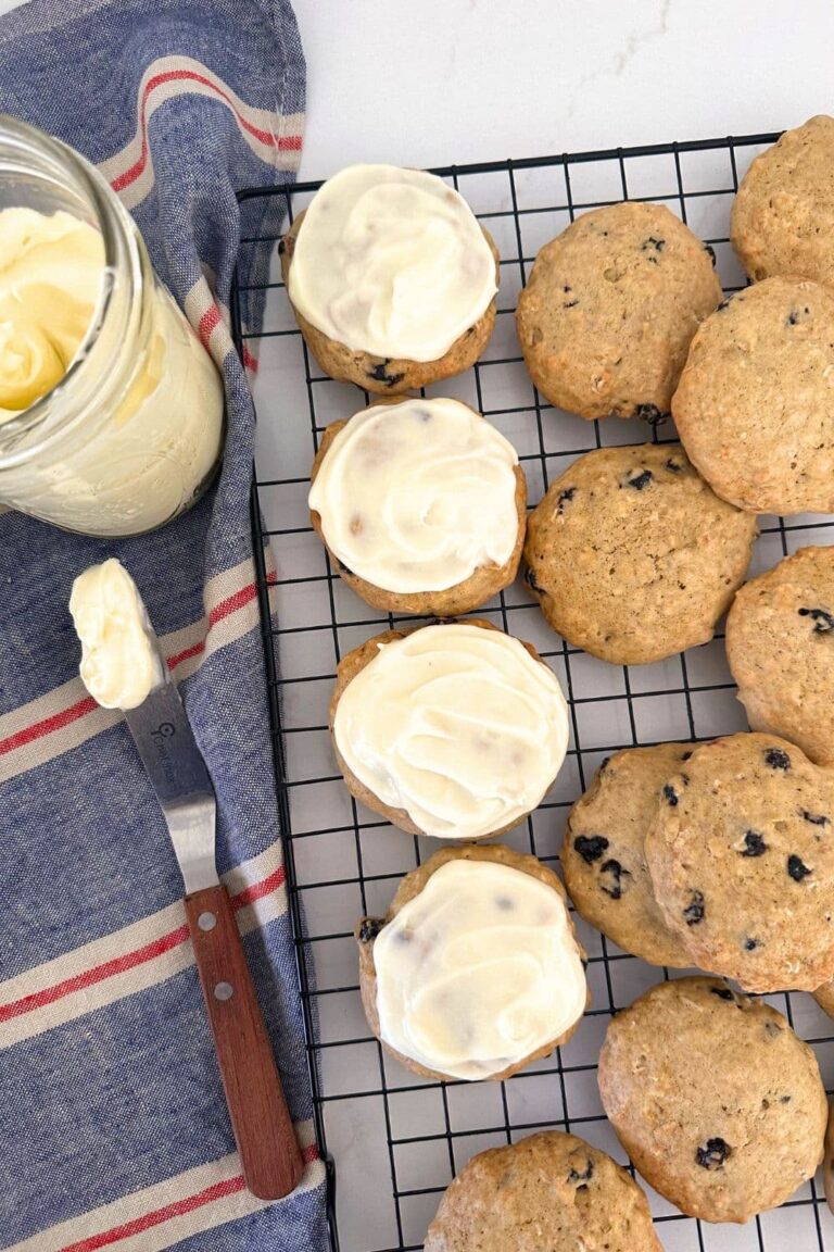 Sourdough blueberry cookies topped with lemon cream cheese frosting sitting on a wire cooling rack. To the left of the cookies you can see a blue and red striped dish towel and a jar of lemon cream cheese frosting with a wooden handled offset spatula.