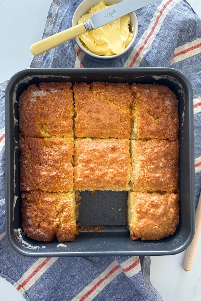 A square cake pan filled with 8 sourdough butter swim biscuits (the 9th biscuit has been removed leaving a hole). There is a red, white and blue dish towel sitting under neath the cake pan.