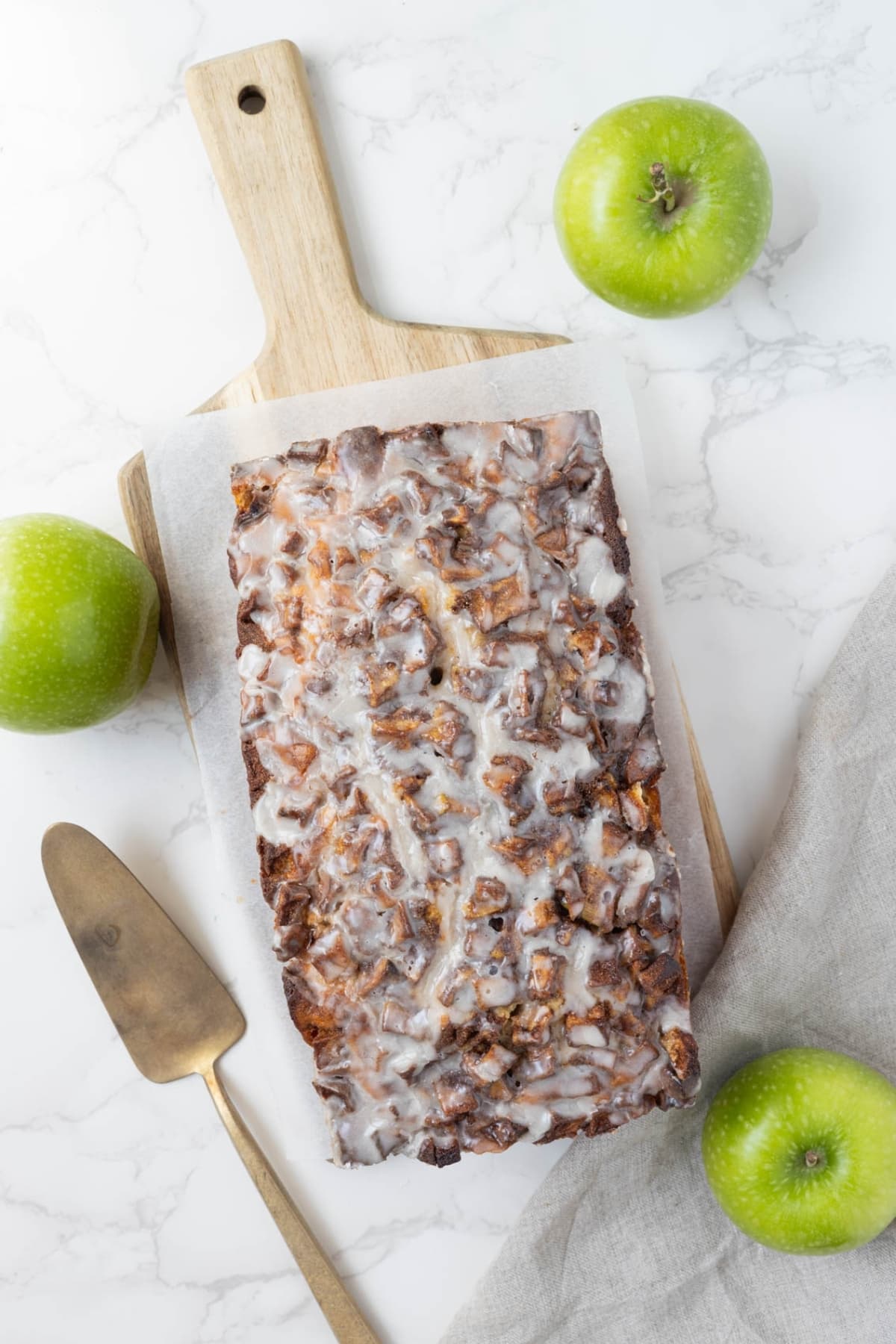 A photo shot top down showing a loaf of sourdough apple fritter bread displayed on a wooden board. There are 3 green apples in the photo as well as a cake slice.
