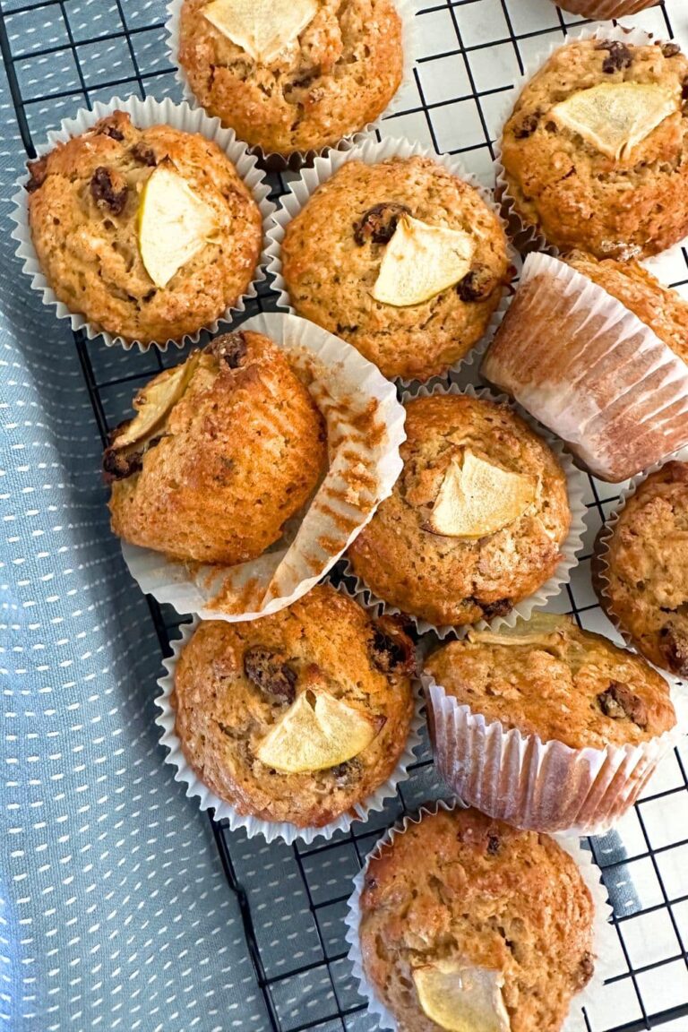 A batch of sourdough applesauce muffins sitting on a black wire cooling rack. There is a light blue dishtowel to the left of the pile of muffins.