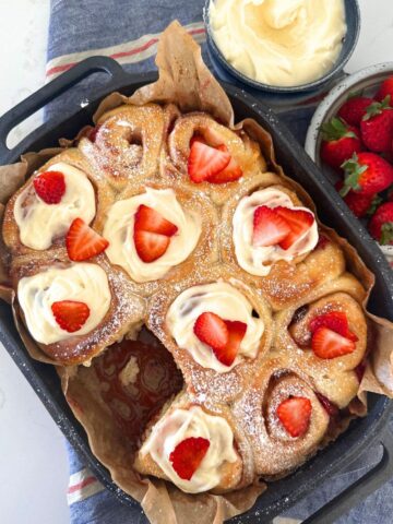 A cast iron baking tray filled with sourdough strawberry rolls topped with cream cheese icing and fresh strawberries.