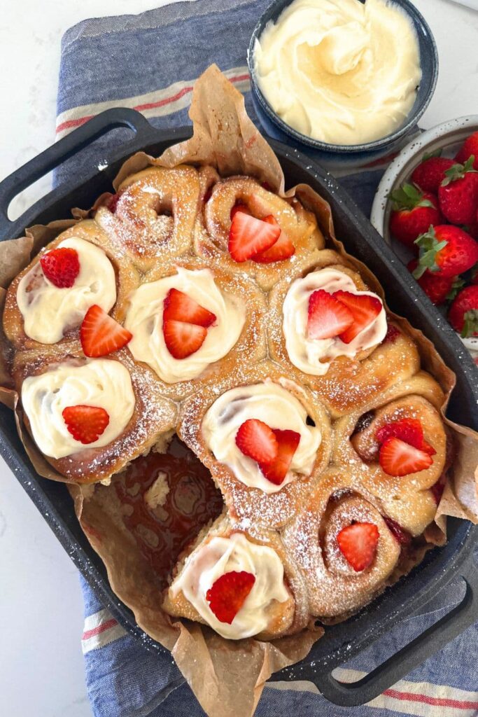 A cast iron baking tray filled with sourdough strawberry rolls topped with cream cheese icing and fresh strawberries.