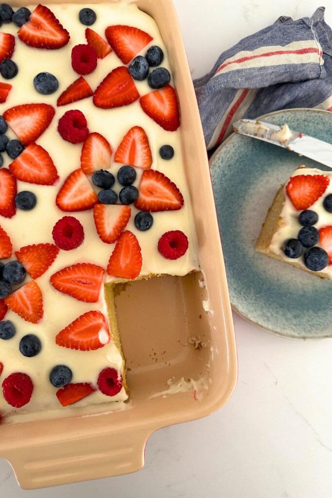 A sourdough vanilla sheet pan cake baked in a baking dish. The cake has been decorated with white frosting and red and blue berries to form red, white and blue decorations. A slice has been taken out of the baking dish and placed on a blue stone ware plate.