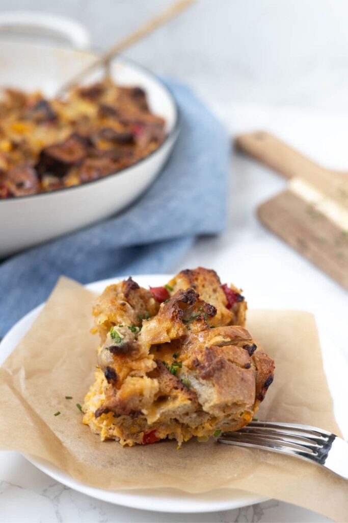 A portion of sourdough breakfast casserole on a white plate with a piece of parchment paper underneath. You can see the skillet of casserole in the background, along with a pale blue dish towel.