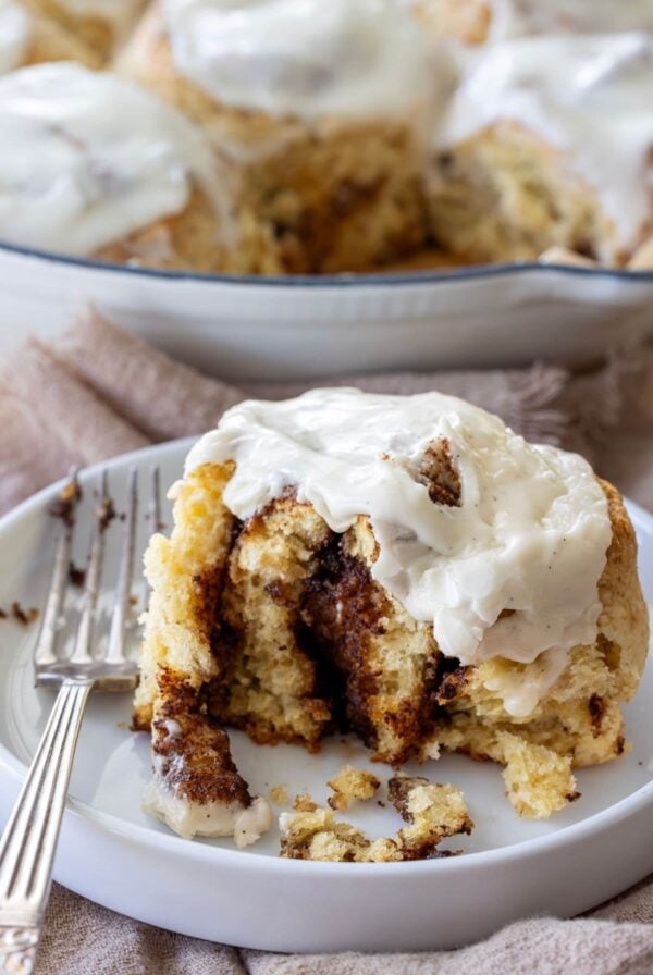 A vertical image showing a cast-iron skillet filled with sourdough discard cinnamon rolls in the background. One of the rolls has been placed onto a white plate in the foreground of the photo.