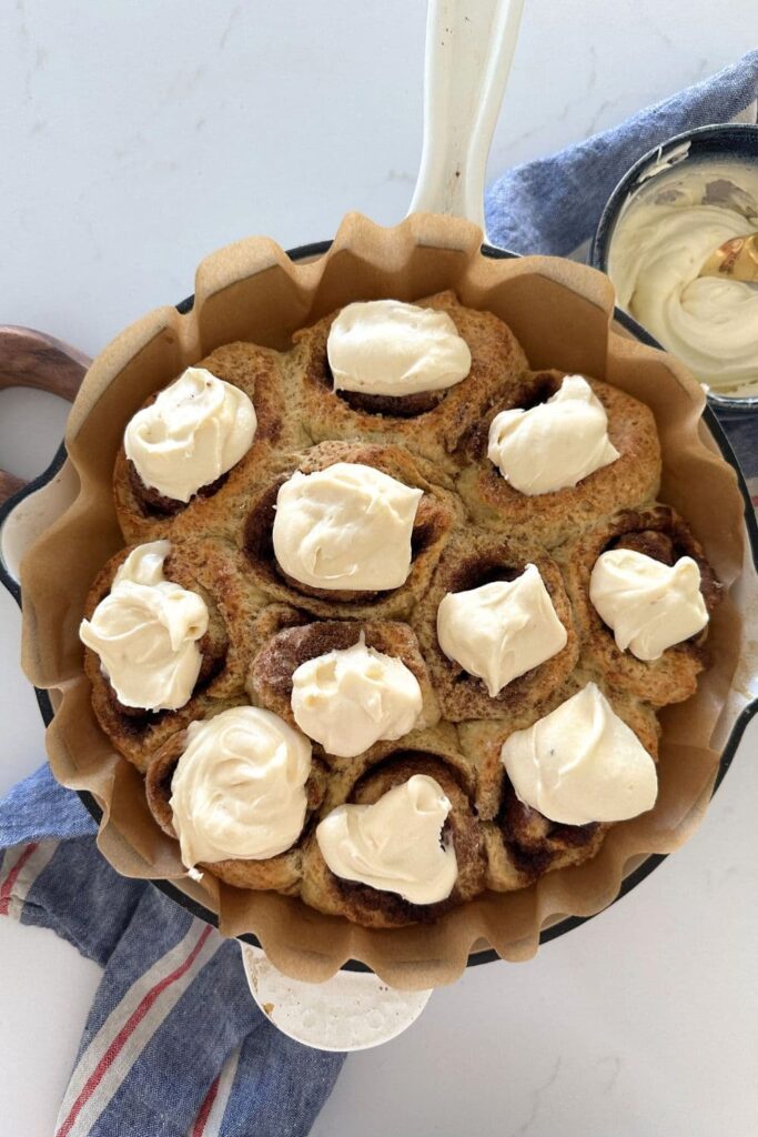 A cream enamel cast iron skillet filled with no wait sourdough cinnamon rolls topped with vanilla cream cheese frosting.

