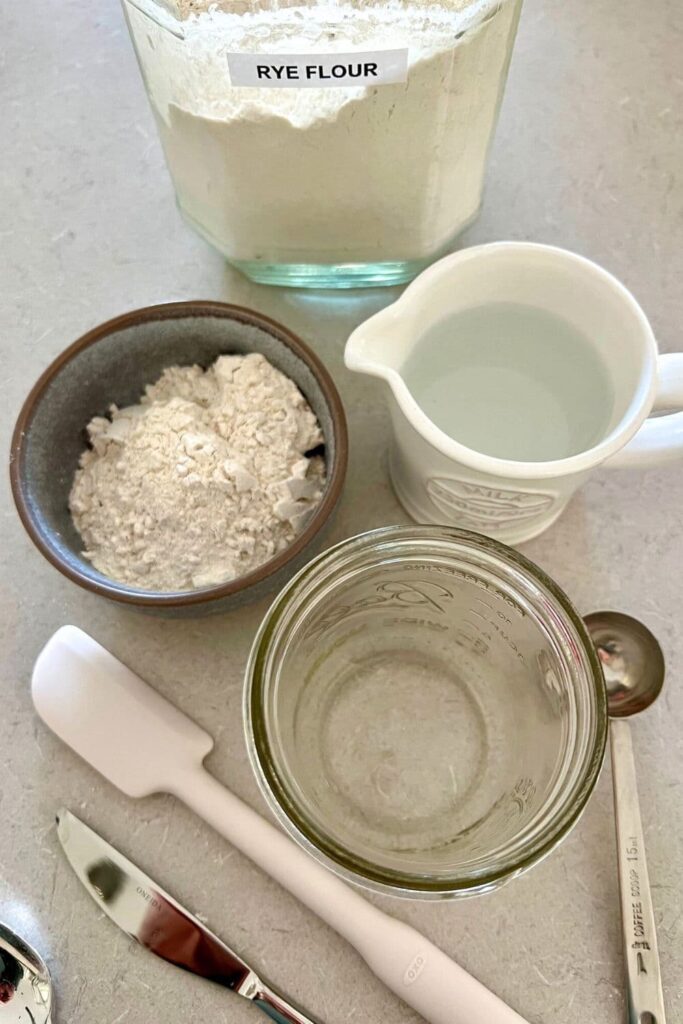 All the tools you need to create a sourdough starter including rye flour, a wide mouth jar, a jug of water, jar spatula and knife for stirring the starter with.