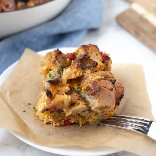 A portion of sourdough breakfast casserole on a white plate with a piece of parchment paper underneath. You can see the skillet of casserole in the background, along with a pale blue dish towel.