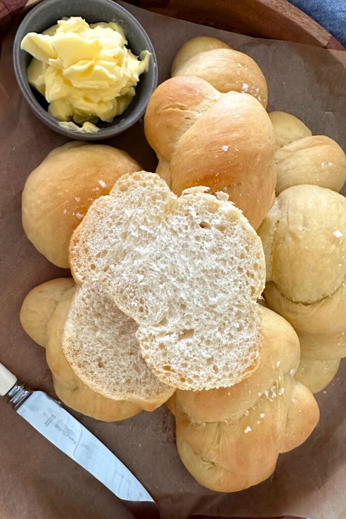 A wooden tray of sourdough buttered knot rolls served with a dish of butter. The roll on the top has been cut open to reveal the soft crumb inside.
