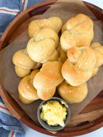 A round wooden tray of sourdough buttered knot rolls displayed with a dish of butter.