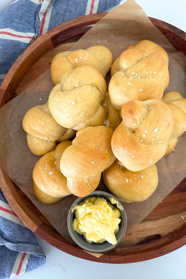 A round wooden tray of sourdough buttered knot rolls displayed with a dish of butter.