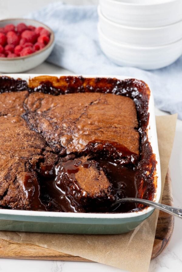 Sourdough chocolate cobbler baked in a square white baking dish. This self saucing pudding is displayed with a bowl of fresh raspberries. There is a spoon that has taken some of the cobbler out to show the rich, chocolate sauce underneath.