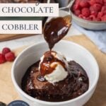 Spoon drizzling fudgy chocolate sauce over sourdough chocolate cobbler topped with vanilla ice cream and text overlay for a Pinterest Pin image.