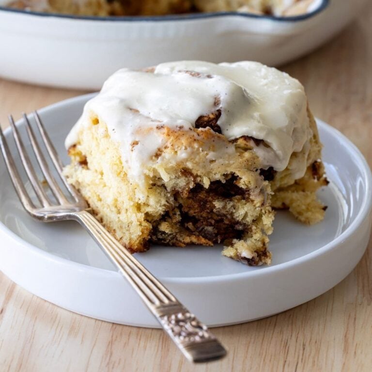Close up image of a sourdough discard cinnamon roll sitting on a white plate. The cinnamon roll is topped with frosting.