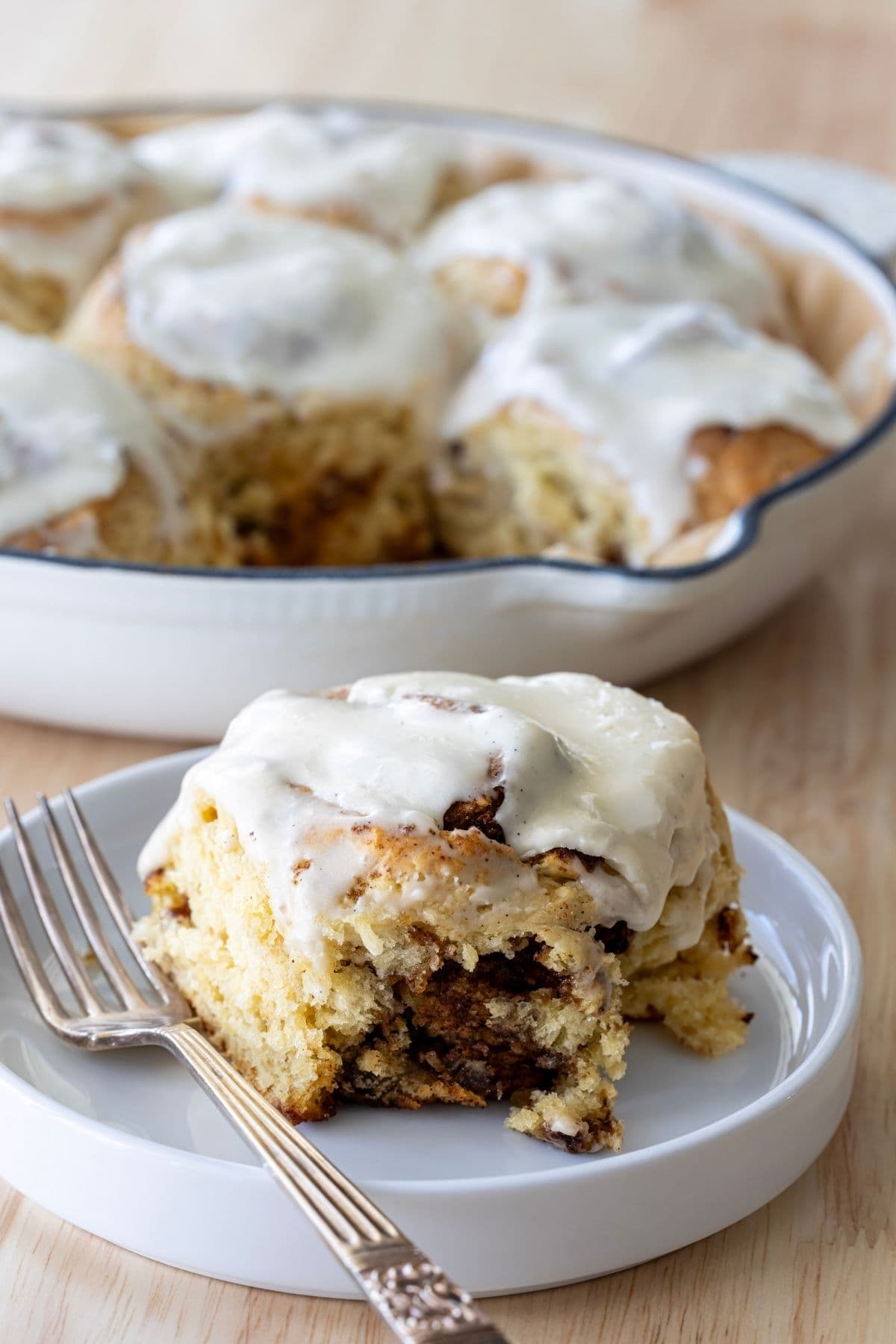 A vertical image showing a cast-iron skillet filled with sourdough discard cinnamon rolls in the background. One of the rolls has been placed onto a white plate in the foreground of the photo.