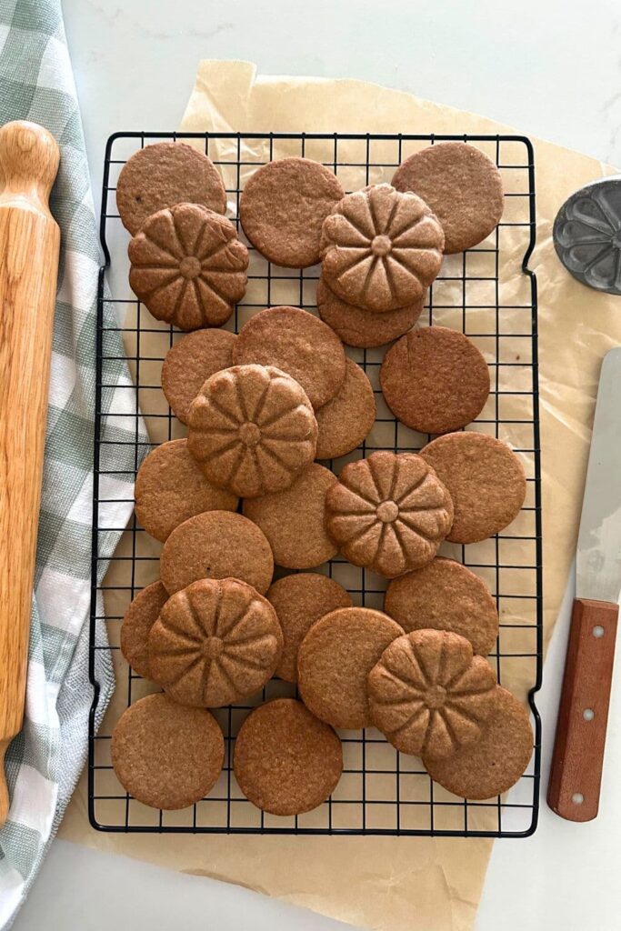 A black wire rack sitting on a piece of parchment paper with a batch of homemade sourdough biscoff cookies sitting on top. There is a checkered dish towel and rolling pin on the left and a wooden handled spatula on the right.