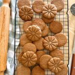 A black wire rack sitting on a piece of parchment paper with a batch of homemade sourdough biscoff cookies sitting on top. There is a checkered dish towel and rolling pin on the left and a wooden handled spatula on the right.