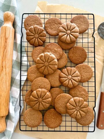 A black wire rack sitting on a piece of parchment paper with a batch of homemade sourdough biscoff cookies sitting on top. There is a checkered dish towel and rolling pin on the left and a wooden handled spatula on the right.