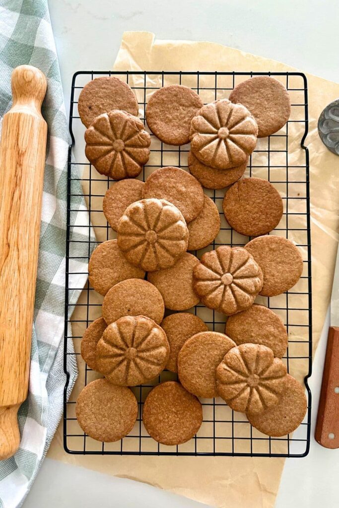 A black wire rack sitting on a piece of parchment paper with a batch of homemade sourdough biscoff cookies sitting on top. There is a checkered dish towel and rolling pin on the left and a wooden handled spatula on the right.