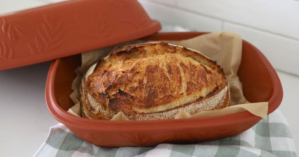 Large loaf of sourdough bread that has been baked in a clay baking dish. It is displayed in the terracotta baking pot with the lid off.