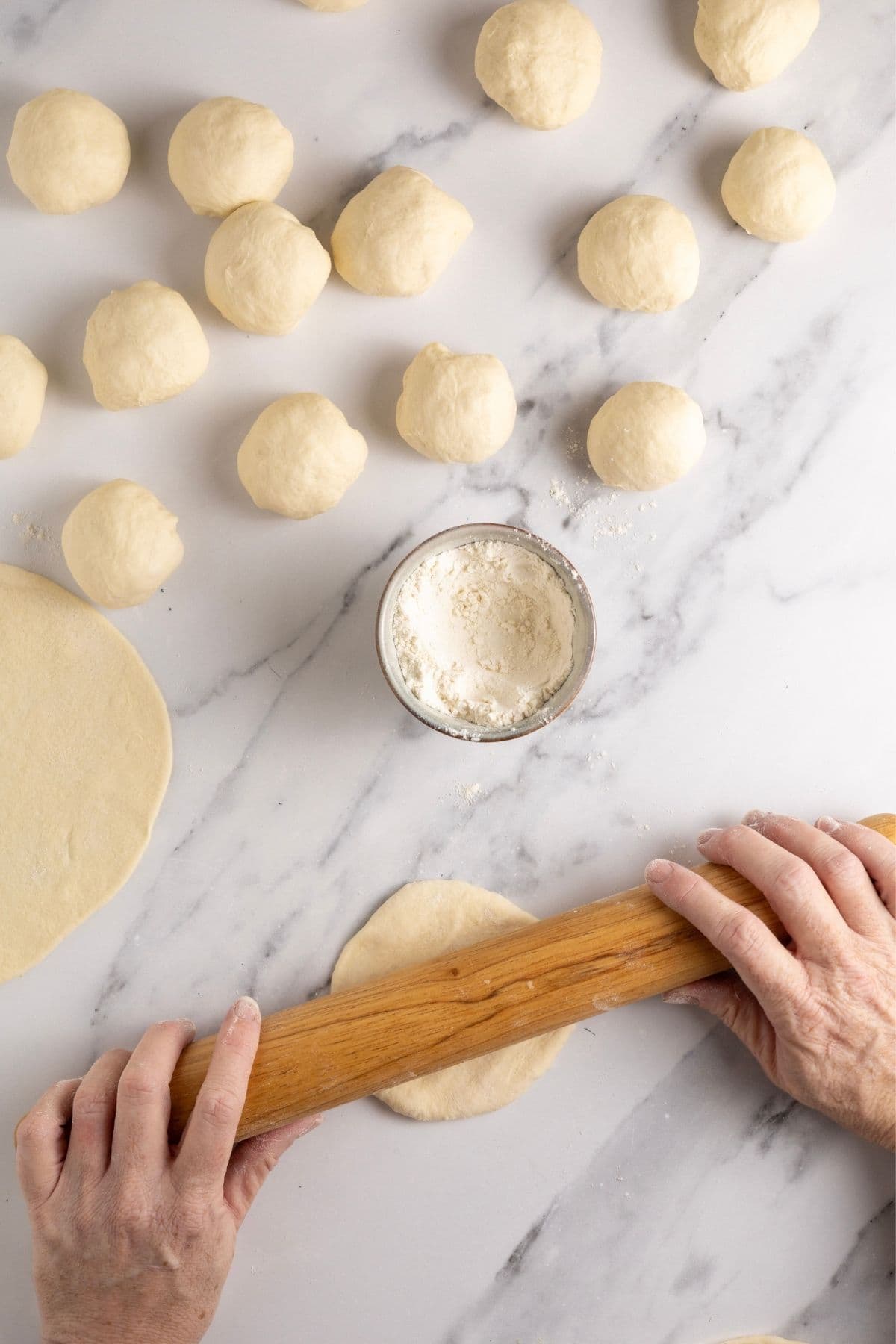 An image showing the dough balls resting on the counter, along with a thin sourdough tortilla that has been rolled out and then a pair of hands holding a rolling pin while rolling out another one of the dough balls.