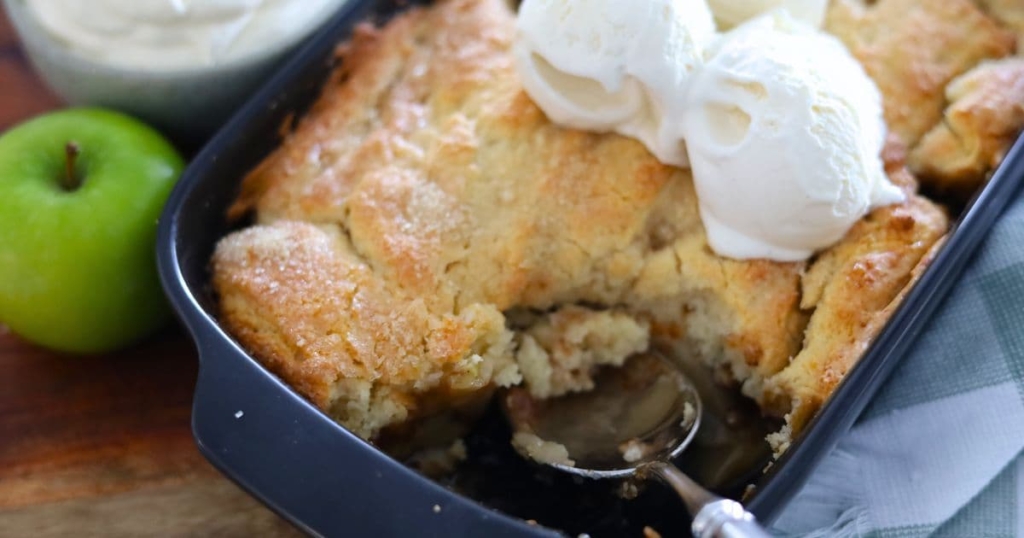 A close up photo of sourdough apple cobbler with a scoop taken out so you can see the soft apple filling underneath. There is vanilla ice cream on top of the cobbler. You can also see a granny smith apple to the left of the baking dish.