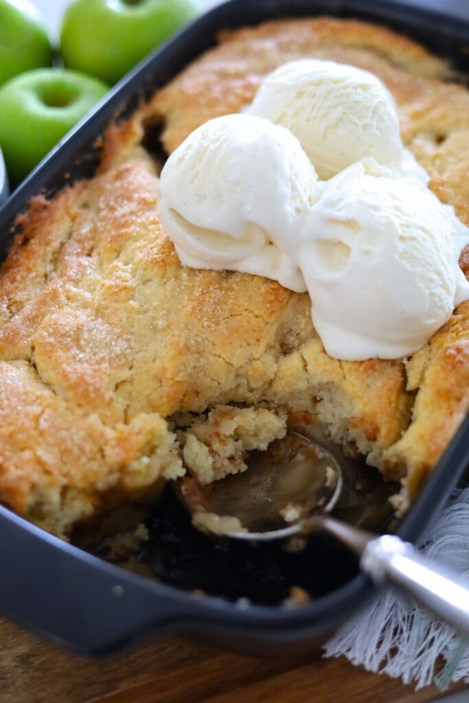 A close up photo of sourdough apple cobbler with a scoop taken out so you can see the soft apple filling underneath. There is vanilla ice cream on top of the cobbler. You can also see a granny smith apple to the left of the baking dish.