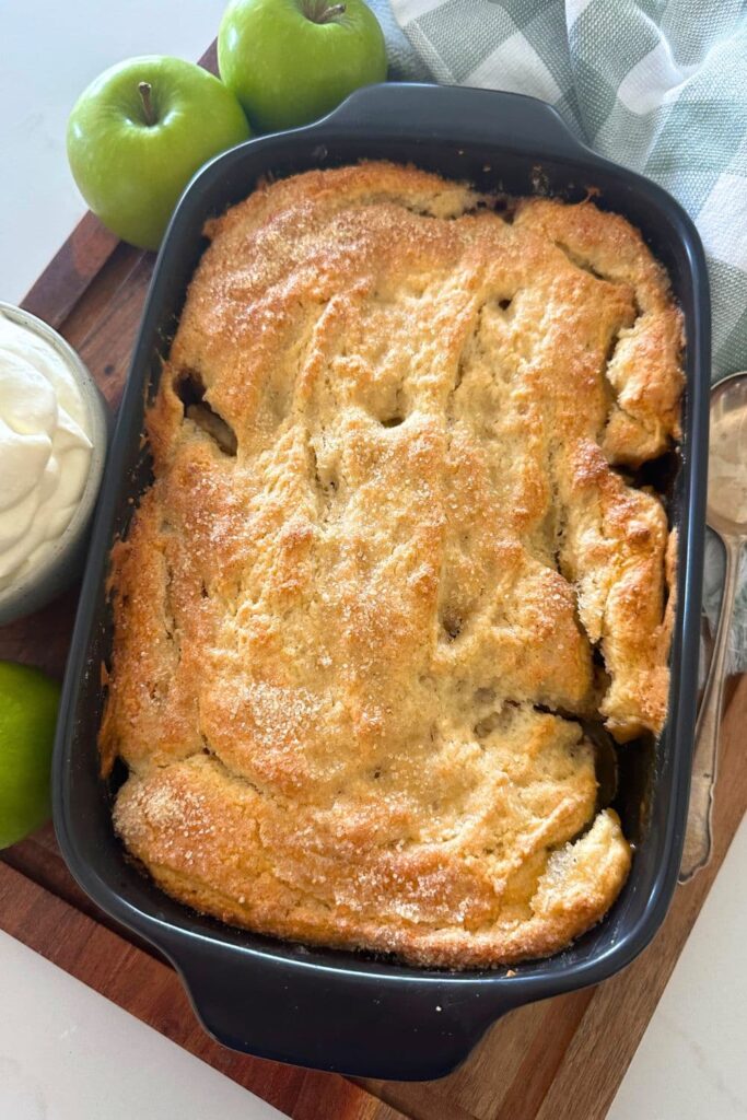 A full length shot of a sourdough apple cobbler in a black baking dish. It has just been taken out of the oven and the topping all golden brown. There are two granny smith apples in the photo too.