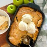 A close up photo of sourdough apple cobbler with a scoop taken out so you can see the soft apple filling underneath. There is vanilla ice cream on top of the cobbler. You can also see a granny smith apple to the left of the baking dish.