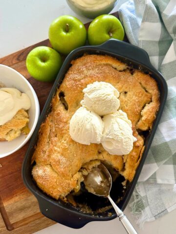 A close up photo of sourdough apple cobbler with a scoop taken out so you can see the soft apple filling underneath. There is vanilla ice cream on top of the cobbler. You can also see a granny smith apple to the left of the baking dish.