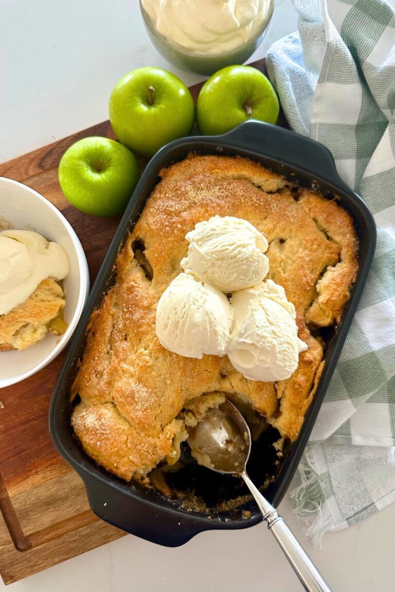 A close up photo of sourdough apple cobbler with a scoop taken out so you can see the soft apple filling underneath. There is vanilla ice cream on top of the cobbler. You can also see a granny smith apple to the left of the baking dish.