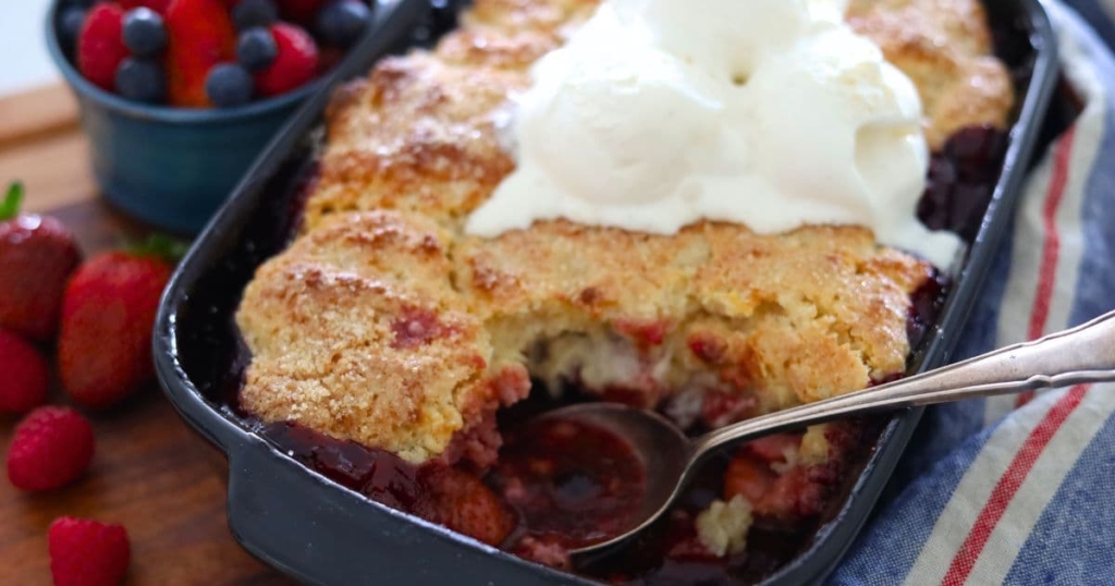 Sourdough berry cobbler baked in a black baking dish. Some of the cobbler has been scooped out so you can see the sweet, jammy filling underneath. The sourdough cobbler is topped with vanilla ice cream that has started to melt.