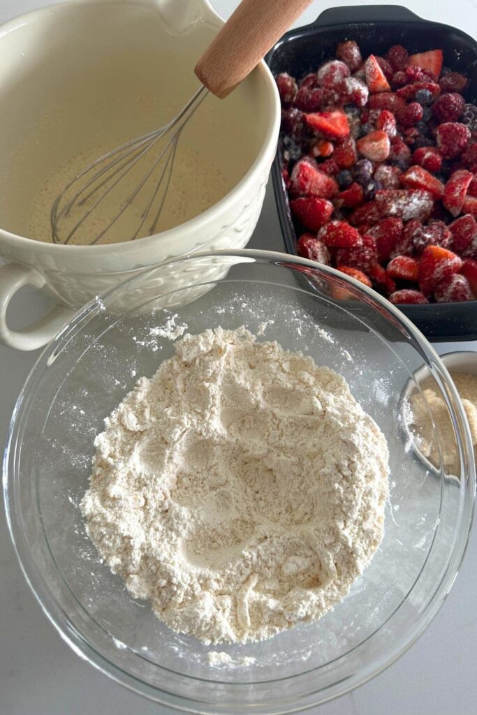 A glass bowl of flour with frozen grated butter rubbed in to it. You can also see liquid ingredients in a cream jug in the background, as well as a black baking dish with the berry filling in it.