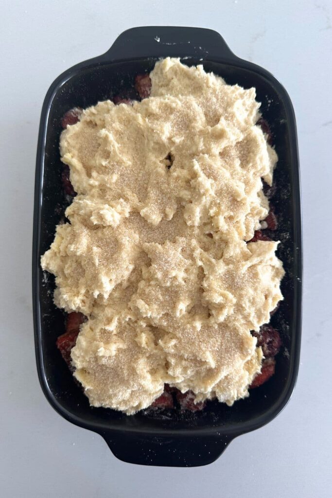 Black baking dish containing sourdough berry cobbler topped with granulated sugar ready to go into the oven.