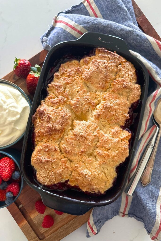 Sourdough berry cobbler baked in a black baking dish. It has just come out of the oven and is displayed on a blue and red striped dish towel with a bowl of whipped cream next to it.