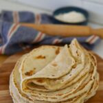 A stack of sourdough tortillas displayed on a round wooden serving board. You can see a wooden rolling pin and bowl of flour in the background.