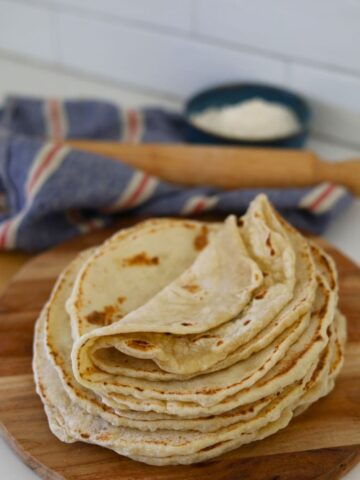 A stack of sourdough tortillas displayed on a round wooden serving board. You can see a wooden rolling pin and bowl of flour in the background.