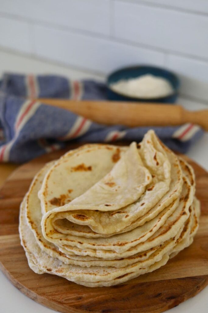 A stack of sourdough tortillas displayed on a round wooden serving board. You can see a wooden rolling pin and bowl of flour in the background.