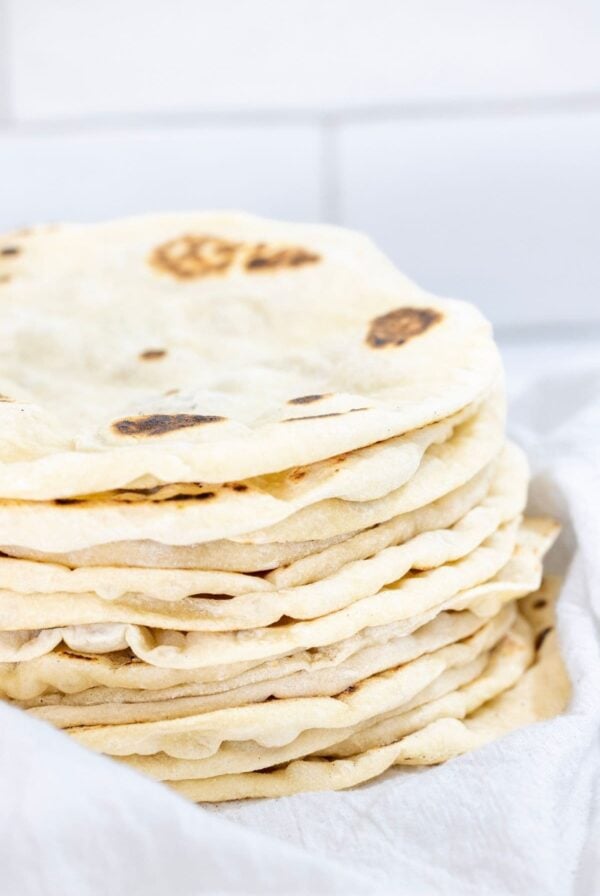 A stack of freshly cooked sourdough discard tortillas nestled in a white flour sack towel. The tortillas are bubbly and have little brown spots on them.