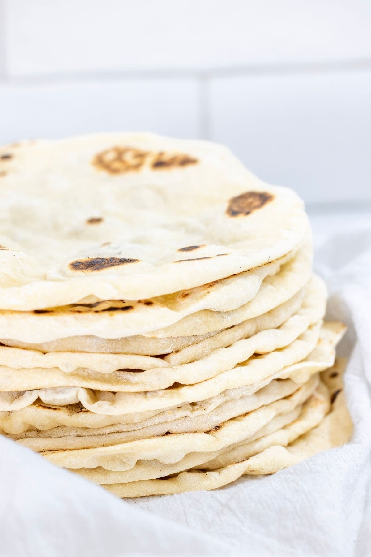 A stack of freshly cooked sourdough discard tortillas nestled in a white flour sack towel. The tortillas are bubbly and have little brown spots on them.