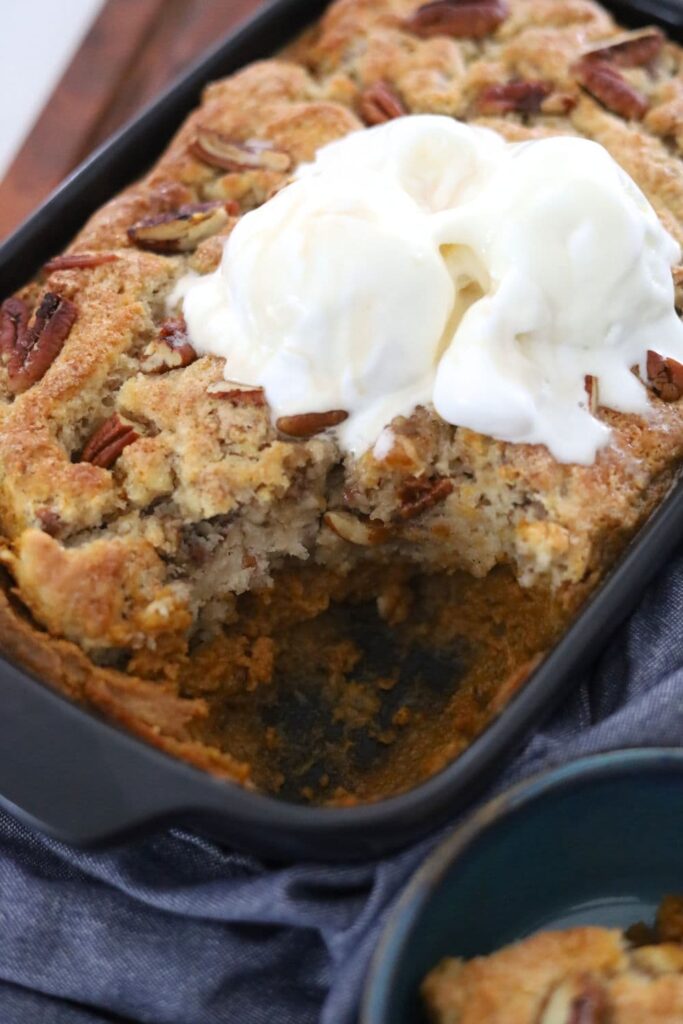 A dark grey baking dish with sourdough pumpkin pecan cobbler inside. There is a scoop missing and you can see the creamy pumpkin filling underneath. There is vanilla ice cream scooped on top of the cobbler.