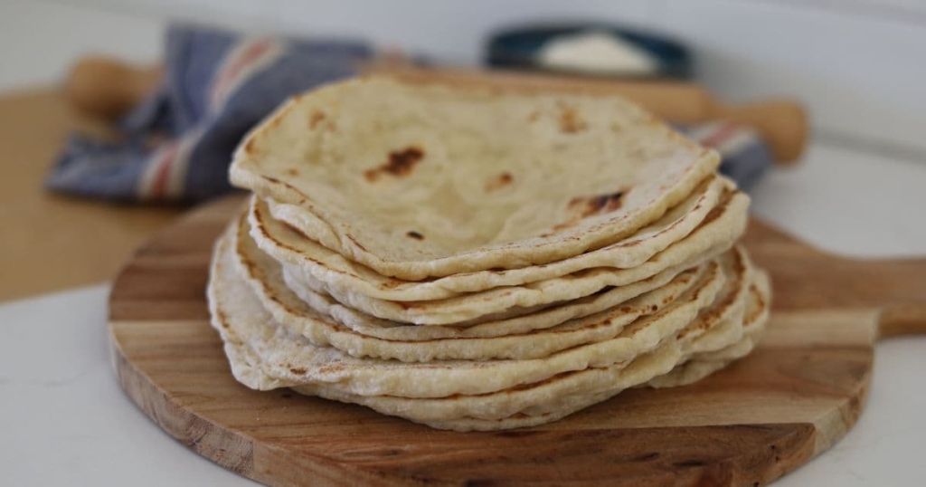 A stack of sourdough tortillas displayed on a round wooden serving board. You can see a wooden rolling pin and bowl of flour in the background.